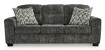 A three-seat sofa is presented in a gray-charcoal fabric with a textured, slightly mottled appearance.  The upholstery has a subtle pattern giving a variation in the shade throughout.  Three matching accent pillows, featuring a pattern of vertical stripes and light beige/cream tones against a darker gray background, are placed on the seat cushions. The sofa has gently curved armrests and dark-colored, faux-wood finish legs. The sofa is presented against a plain white background, which helps to highlight the sofa's color and texture.