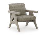 A single, stylish outdoor armchair photographed against a plain, white background. The chair has a modern, slightly rustic design that combines a light wood frame with a woven seat and backrest. The wooden frame appears to be made from a pale, natural-toned wood with a smooth, matte finish. Its structure is sturdy and geometric, featuring angled legs that splay outward slightly for stability. The armrests are thick and straight, extending horizontally from the back toward the front. Each armrest is supported by a distinctive triangular side structure formed by the legs and cross supports, giving the chair a sculptural, architectural look. The front legs angle forward while the rear legs angle slightly backward, creating a balanced stance. The seat and backrest are made of tightly woven material in a neutral beige or taupe color, resembling wicker or rattan. The weaving pattern is consistent and textured, adding visual interest and a tactile quality. The backrest is slightly reclined, suggesting comfort, while the seat is firm and gently contoured. Small, dark protective caps are visible at the bottom of each leg, likely to prevent floor damage. Overall, the chair has a clean, contemporary aesthetic with natural materials and a neutral color palette, making it suitable for both indoor and outdoor settings.